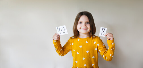 cute kid girl in mustard dress learns the multiplication table on beige background, child with an example sheet and numbers in his hands