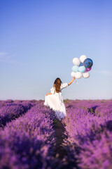 girl in a long white dress walks in a field of lavender. View from the back. In the hands of a large bunch of balloons