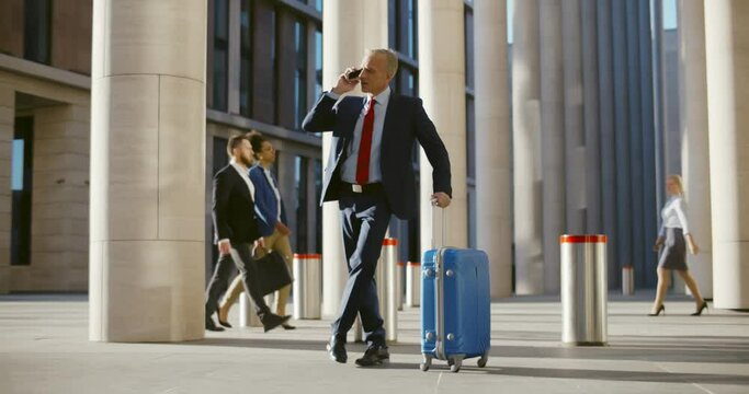 Happy mature businessman chatting on phone arriving at airport with suitcase standing outside.