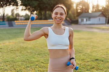 Outdoor shot of young female athlete with dumbbells stretching and warming up in stadium, looking smiling at camera, expressing happiness, sport and fitness.