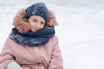 A teenage girl in a snowy blue hat and a pink jacket sits in the snow and smiles. People, lifestyle concept
