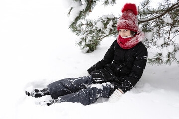 A teenage girl in a snow-covered red hat and a black jacket sits in the snow under a pine tree and smiles. People, lifestyle concept