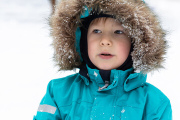 A boy in a snow-covered turquoise winter jacket with a hood on a white background looks into the distance. People, lifestyle concept