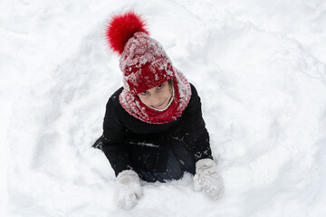 A teenage girl in a snowy red hat and a black jacket sits in the snow and smiles. People, lifestyle concept