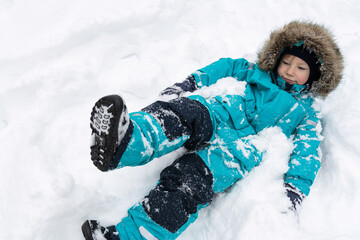 A boy in a snow-covered turquoise winter overalls with a hood lies in the snow and smiles. People, lifestyle concept