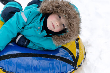 A boy in a turquoise overalls with a hood lies on a tubing in the snow and smiles. People, lifestyle concept