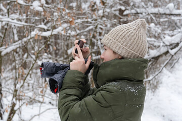 A teenage girl in a beige hat and an olive-colored jacket stands in a snowy forest under a snowfall and takes pictures on the phone. People, lifestyle concept