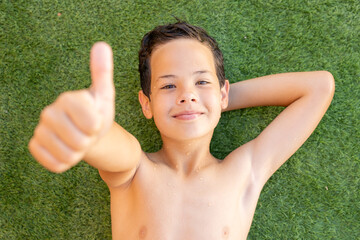 Happy boy lying down on the grass with thumb up. Summer season.