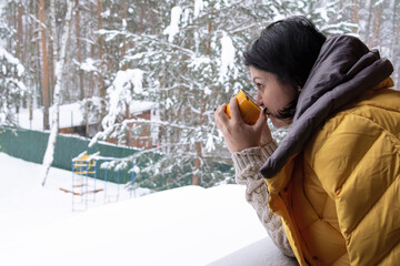 A woman in a yellow down jacket stands on the terrace of a house and drinks coffee from a yellow cup against the backdrop of a snow-covered forest