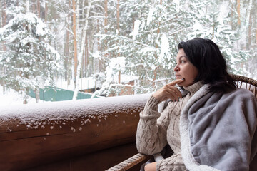 A woman in a beige sweater and a gray plaid sits on an armchair on the balcony of a building and looks at the snow-covered forest