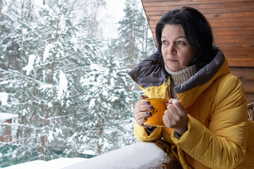 A woman in a yellow jacket and with a yellow cup with coffee in her hands stands on the terrace of a house and looks into the distance against the background of a snow-covered forest