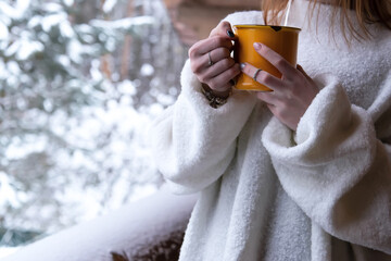 A woman in a white sweater and a yellow cup with coffee in her hands is standing on the terrace of a house against the backdrop of a snow-covered forest