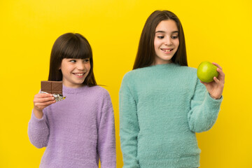 Little sisters isolated on yellow background taking a chocolate tablet in one hand and an apple in...