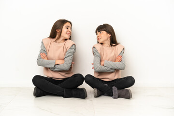 Little sisters sitting on the floor isolated on white background looking over the shoulder with a smile