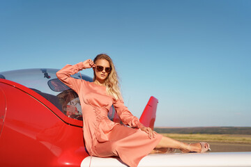 Pretty blond woman with wavy hair wearing pink silk classy dress posing near a private plane