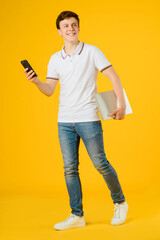 Portrait of a young happy man in white t-shirt smiling and holding a notebook computer  and a smartphone on a yellow background.