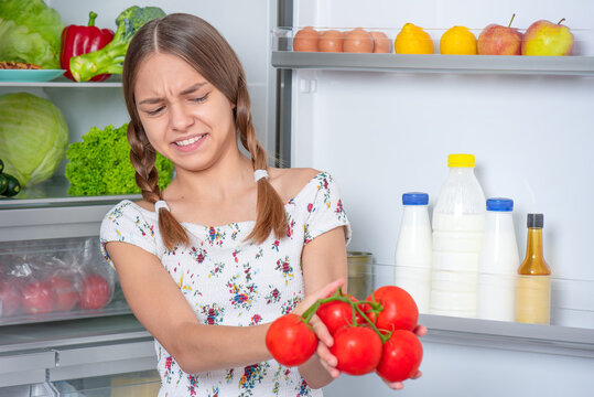 Smiling beautiful young teen girl holding fresh red tomatoes while standing near open fridge in kitchen at home. Portrait of pretty child choosing food in refrigerator full of healthy products