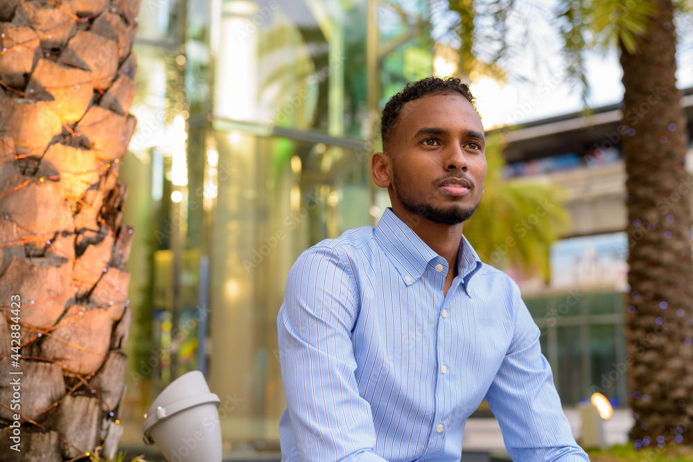 Wall mural Portrait of handsome black African businessman outdoors in city during summer sitting and thinking