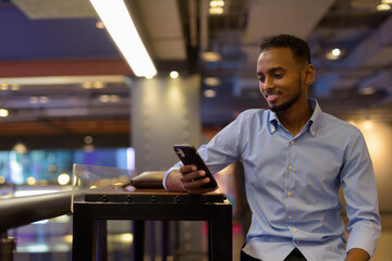 Portrait of handsome black African businessman sitting inside shopping mall smiling and using phone