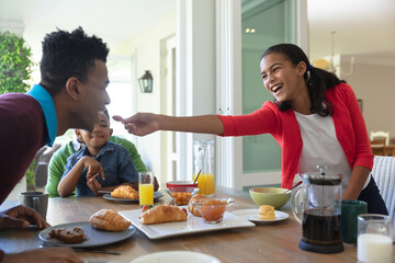 Happy african american son and father sitting at table during breakfast, daughter feeding him