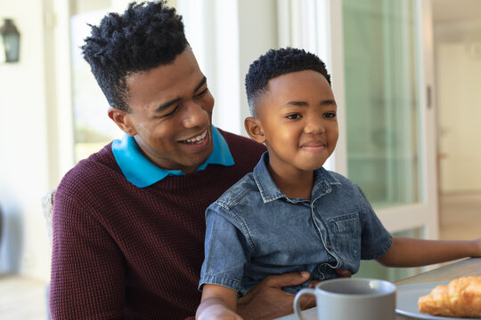 Happy african american son sitting on father's knee at table during breakfast and smiling
