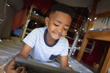 Happy african american boy at home, lying in blanket fort and using tablet - Powered by Adobe