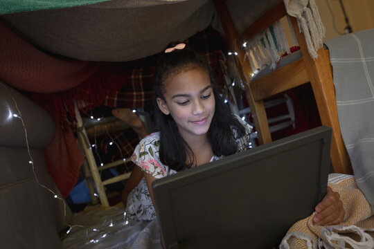 Happy african american girl at home, lying in blanket fort and using tablet