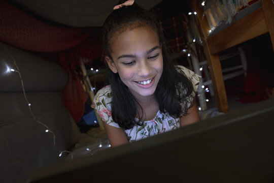 Happy african american girl at home, lying in blanket fort and using tablet