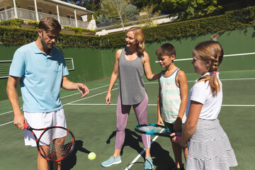 Happy caucasian couple with daughter and son outdoors, playing tennis on tennis court