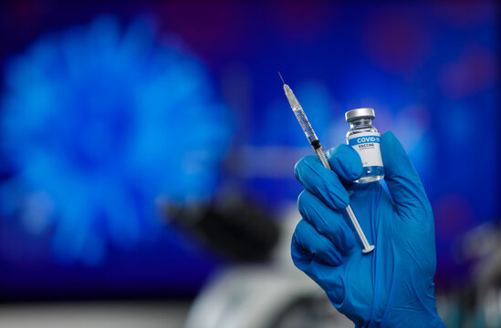 Hand wearing blue medical gloves hoding bottle of Covid-19 vaccine and syringe ready to give injection with virus image blur in background