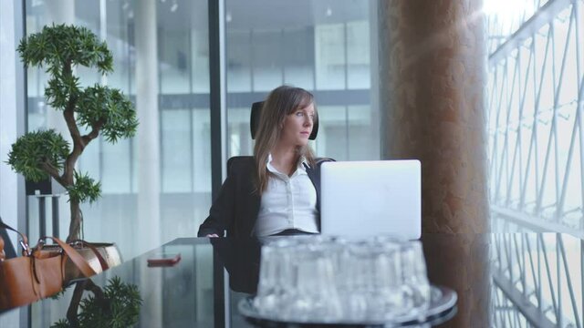 Businesswoman working at desk with laptop computer in corporate office, thinking, waiting for partners in meeting room. 4K video footage.