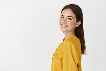 Young confident woman turn head at camera, looking happy and smiling, standing over white background in yellow blouse