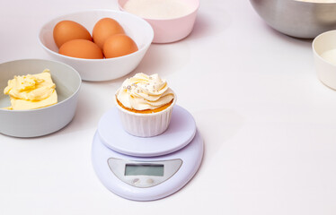 Freshly prepared cupcake with butter cream on the scales, there are ingredients for its preparation next to it. Top view. White background with space for text. Selective focus.
