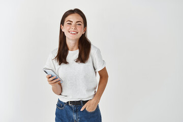 Image of smiling woman using mobile phone and looking at camera happy, standing over white...