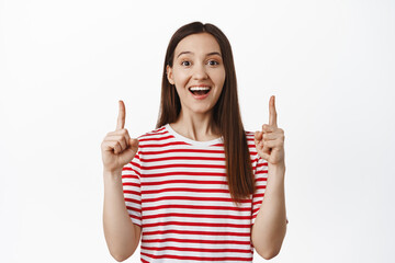 Big announcement on top. Excited smiling girl pointing fingers up happy, showing sale advertisement, logo or banner upwards, standing against white background