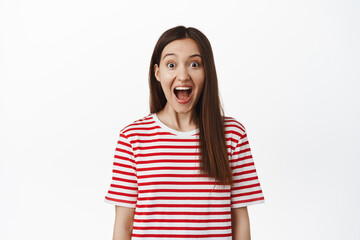 Image of excited surprised girl drop jaw, scream from amazement and looking at camera amazed, staring in awe at surprise for her, standing in striped t-shirt against white background