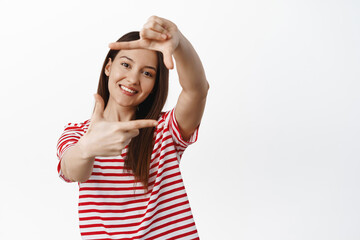 Obraz na płótnie Canvas Portrait of cute smiling girl looking through hand frames camera gesture, picturing moment, taking photo of smth, standing in striped t-shirt against white background