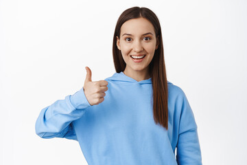 Very good. Image of young woman in hoodie showing thumb up in approval, like and agree, praise good work, satisfied with quality, recommend smth, white background