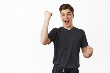 Fototapeta premium Success. Smiling happy man fist pump, say yes and look excited, winning and celebrating, triumphing from win, enjoy taste of victory, chanting, standing over white background