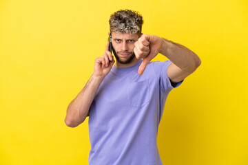 Young caucasian man using mobile phone isolated on yellow background showing thumb down with negative expression