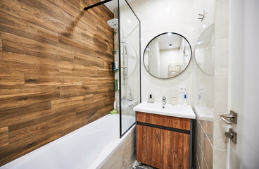 Stylish and comfortable version of bathroom design in small space. White color of rectangular bathtub and wash basin in harmonious combination with round mirror supplemented by wooden decor. - obrazy, fototapety, plakaty