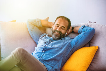 Middle aged man listening music with headphones relaxed in sofa at his home. Middle-aged man relaxing at home with his music listening to tunes on his mobile phone using an ear bud with closed eyes. 