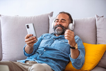 Middle aged man listening music with headphones relaxed in sofa at his home. Cropped shot of a...