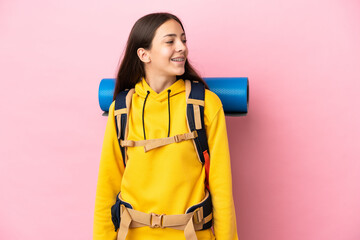 Young mountaineer girl with a big backpack isolated on pink background looking side