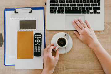 Top view of female manicured hands in an office, wooden desk table with laptop notebook, remote contol, notebook and cup of coffee.