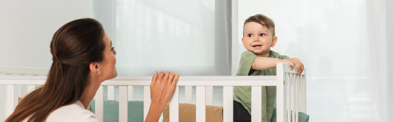 Woman standing near smiling kid in baby bed, banner