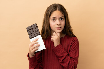 Little caucasian girl isolated on beige background taking a chocolate tablet and having doubts
