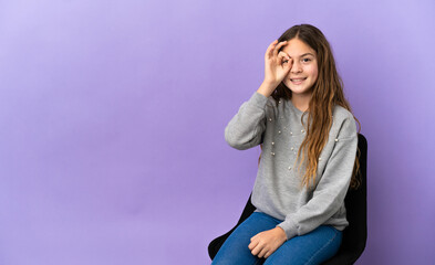 Little caucasian girl sitting on a chair isolated on purple background showing ok sign with fingers