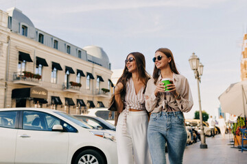 Beautiful stylish young girls in great mood laughing and walking in city center. Women in blouses enjoys sunny summer day and holds coffee