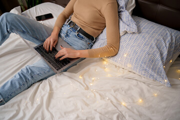 Cropped image of woman on white bed in jeans with a laptop computer. Concept of happy living, relaxation, comfort, stay home, internet surfing and communicating